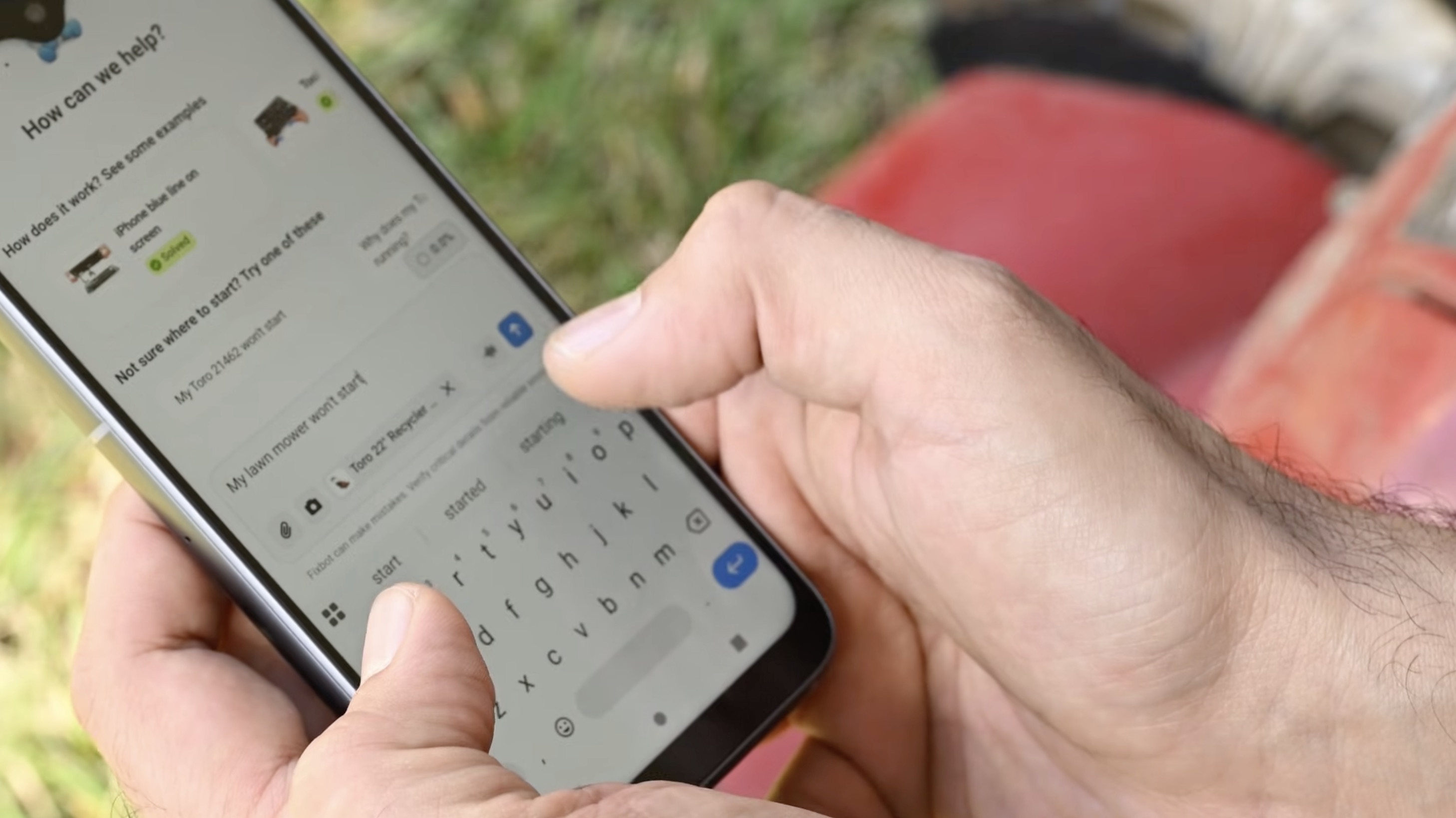 A person holds a smartphone outdoors and types a troubleshooting question about a lawn mower into a chat interface, with grass and a red lawn mower visible in the blurred background 