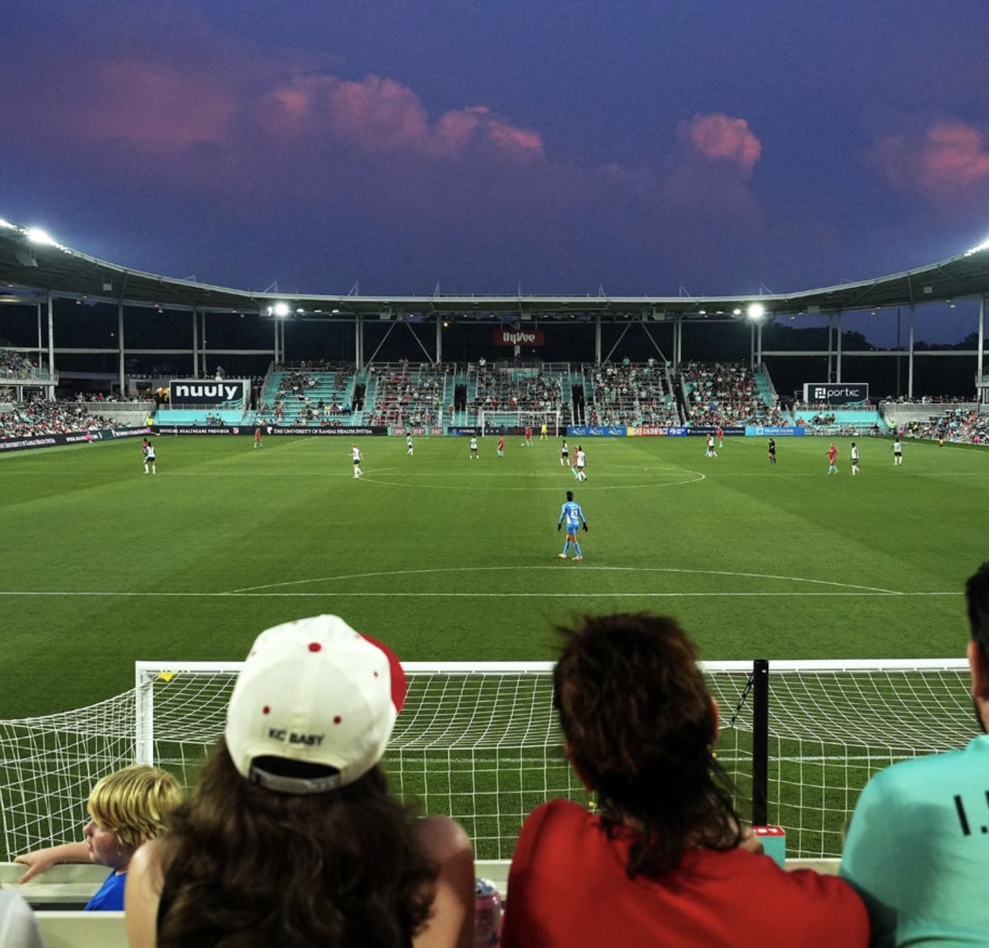 Evening match at CPKC Stadium with fans seated behind a goal, watching two women’s teams play on a brightly lit field. The stands are nearly full, and a purple-tinted sky hangs over the world’s first purpose-built venue for a professional women’s sports team 